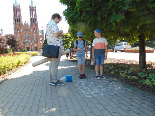 Dwoje dzieci i mężczyzna stoją na chodniku z namalowanym kredą prostokątem zawierającym litery. W tle kościół w stylu neogotyckim, wybudowany z czerwonej cegły