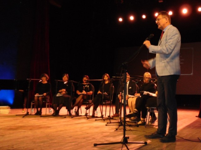 Drewniana scena, reflektory, na krzesłach 9 osób ubranych na czarno, mężczyzna stoi przy mikrofonie