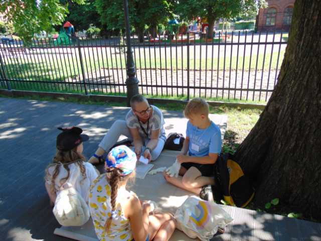 Grupka dzieci siedzi pod drzewem i w jednorazowych rękawiczkach oglądają starą książkę