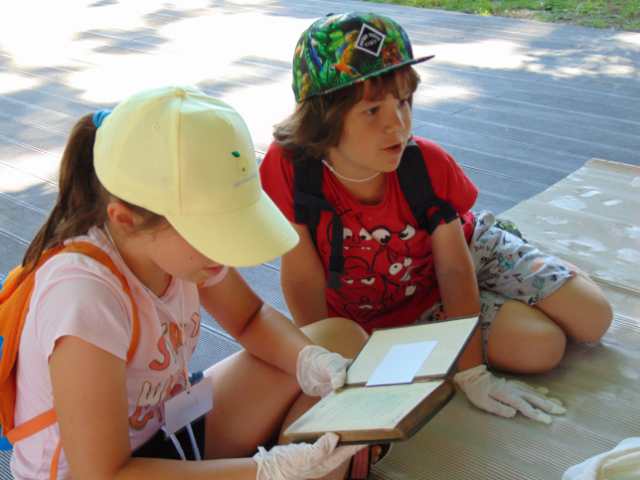 Chłopiec i dziewczynka w jednorazowych rękawiczkach siedzą na tekturze falistej. Dziewczynka czyta starą książkę