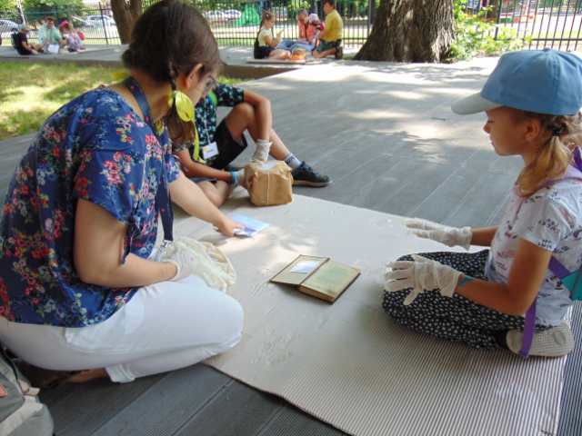 Dwoje dzieci i opiekunka siedzą na deskach, na tekturze falistej, między nimi stara książka i paczka zapakowana w szary papier i przewiązana sznurkiem