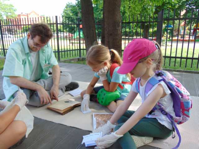 Dwoje dzieci i dorosły opiekun siedzą na podeście z desek, na tekturze falistej, oglądają starą książkę