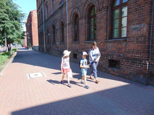 Dwoje dzieci i kobieta idą szerokim chodnikiem, w tle dwie kamienice z czerwonej cegły