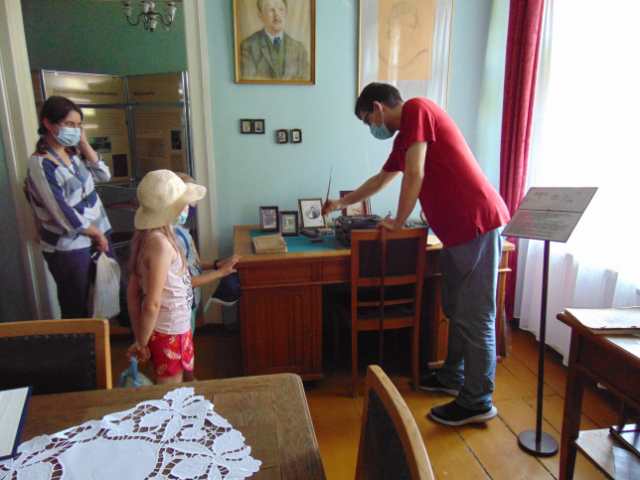 Pokój, w nim dwoje dzieci i dwoje dorosłych. Stare drewniane biurko. Mężczyzna w czerwonej koszulce pokazuje jak pisało się prawdziwym gęsim piórem